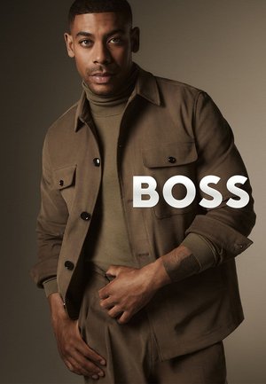 Bruine stoffen jas met zakken met knopen, gedragen over een groene coltrui. Model poseert zelfverzekerd tegen een neutrale achtergrond. Tekst "BOSS" zichtbaar.