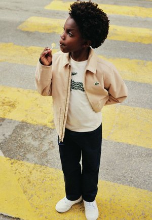 Beige Jacke, weisses T-Shirt mit grünem Motiv, dunkle Hosen und weisse Turnschuhe. Kind hält einen Lollipop und steht auf einem gelben Zebrastreifen.