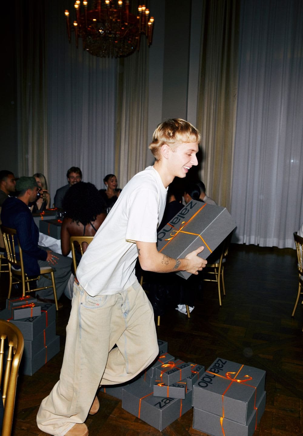 Grå gaveæsker med orange bånd er stablet på gulvet. En person iført hvid T-shirt og beige bukser bærer en kasse.