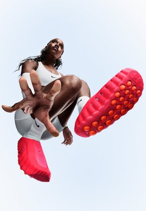 Zapatillas Nike con parte superior de color rosa brillante, superficie texturizada y detalles anaranjados en la suela. Modelo en movimiento, vestido con un conjunto blanco.