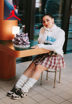 Chemise white surdimensionnée avec "Jordan" en teal, jupe en tartan, chaussettes blanches montantes, et sneakers noir et blanc. Dessert sculpté sur la table.