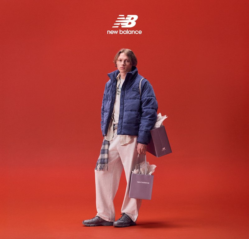 Giacca trapuntata blu, pantaloni grigio chiaro, scarpe grigie. Tiene borse della spesa su sfondo rosso. Sciarpa a quadri e camicia a strati.