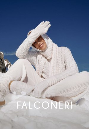 Persona vestita con maglieria bianca, inclusi guanti, sciarpa e cappuccio, seduta sulla neve con cielo azzurro limpido e cime montuose sullo sfondo.