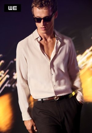 Hvid skjorte med knapper og afslappet pasform, parret med mørke bukser og et struktureret bælte. Solbrillerne forbedrer det moderne, elegante udseende.
