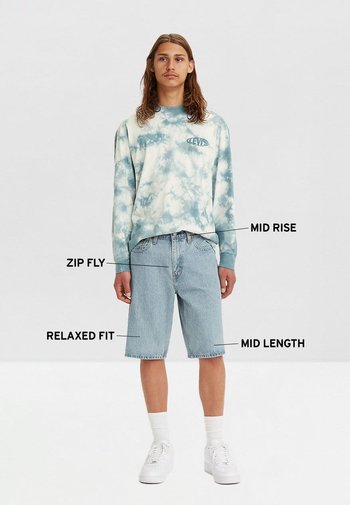 Sudadera azul claro con diseño de 'tie-dye' y un logo, combinada con pantalones cortos de mezclilla de tiro medio y corte relajado, y zapatillas blancas con calcetines tobilleros.