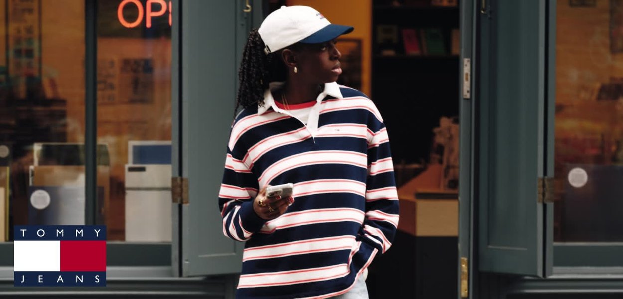 Giovane persona con indosso una maglietta a righe Tommy Jeans e un cappello bianco, che tiene in mano uno smartphone, in piedi fuori da un negozio con le porte aperte.