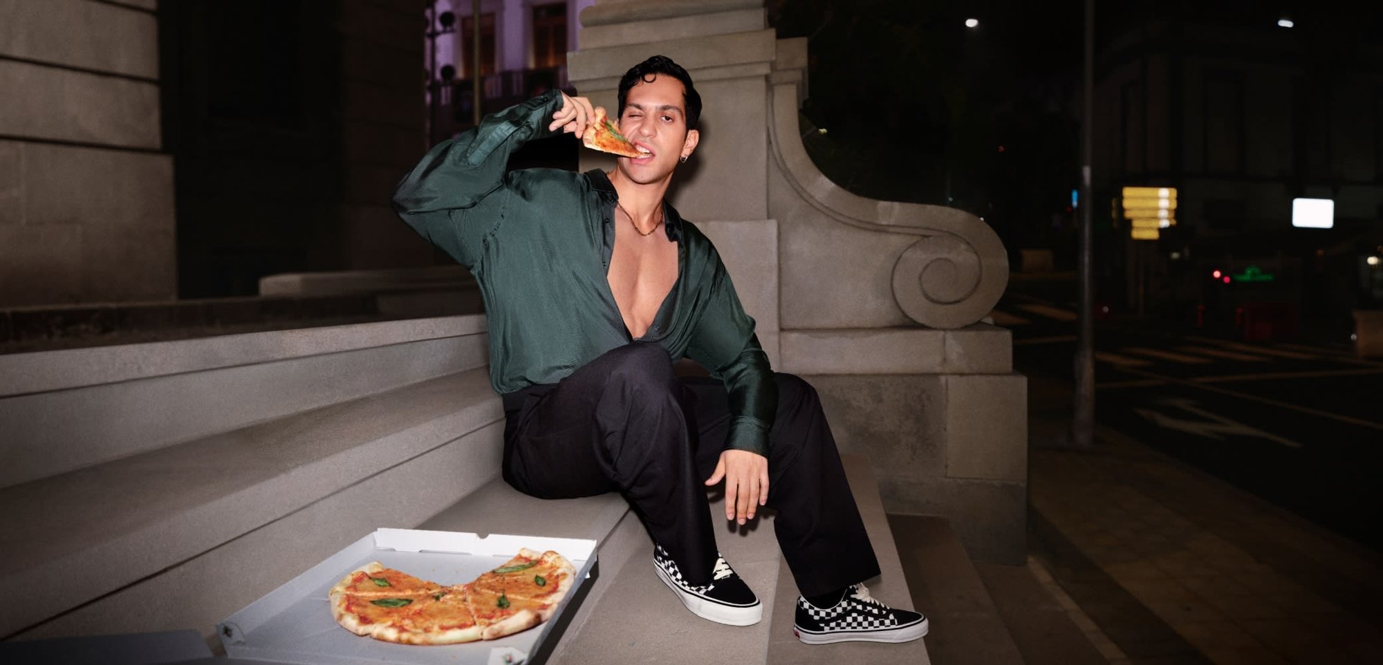 Una persona con una camiseta de color verde oscuro y pantalones negros se sienta en unas escaleras de piedra, sosteniendo una porción de pizza. Una caja de pizza descansa cerca. Se pueden ver zapatos de cuadros.