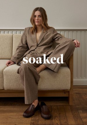 Frau mit langen Haaren trägt einen braunen Nadelstreifen-Anzug und liegt entspannt auf einem beigen Sofa, mit Loafers auf dem Holzboden.