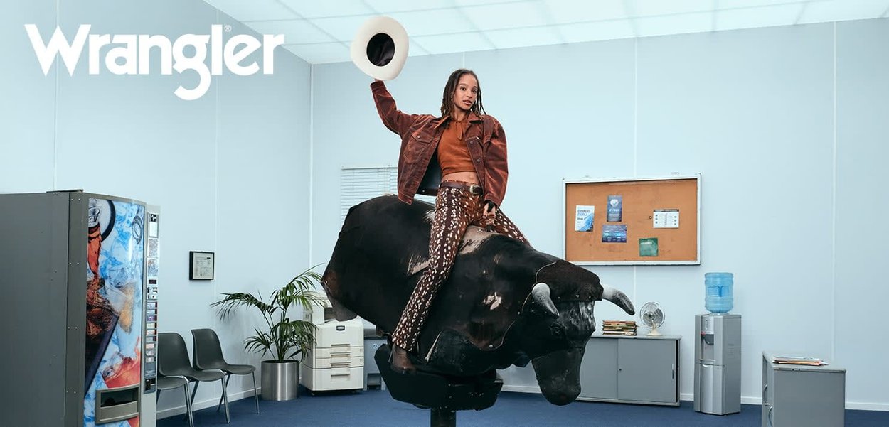 Wrangler brown leather jacket over an orange top, patterned pants, and white hat, riding a black bull statue in an office setting.