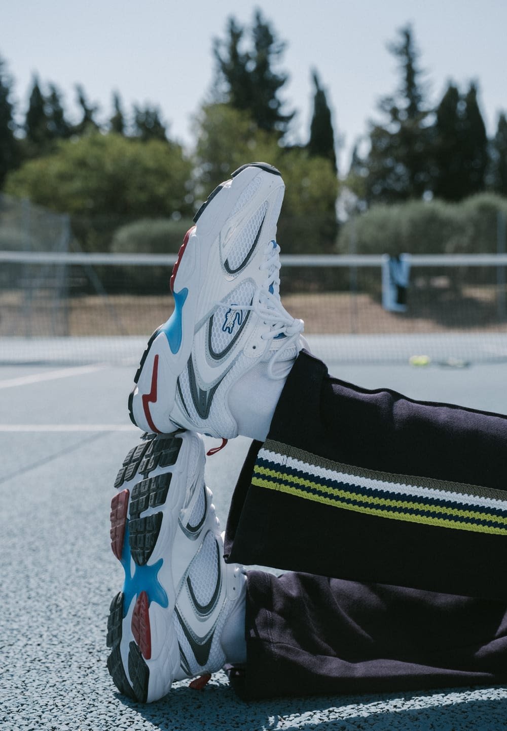 Weiße Sportschuhe mit blauen, roten und grauen Akzenten, ausgestattet mit einem Mesh-Obermaterial, strukturierten Sohlen und einem auffälligen Schnürsystem, das auf einem Platz liegt.