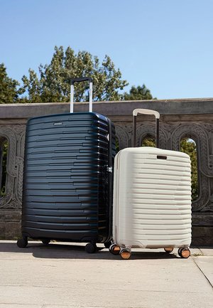 Neben einander stehen zwei Koffer: ein dunkelblauer, gerillter Hartschalenkoffer und eine weiße, kompakte Version, beide mit ausziehbaren Griffen und Rollen.
