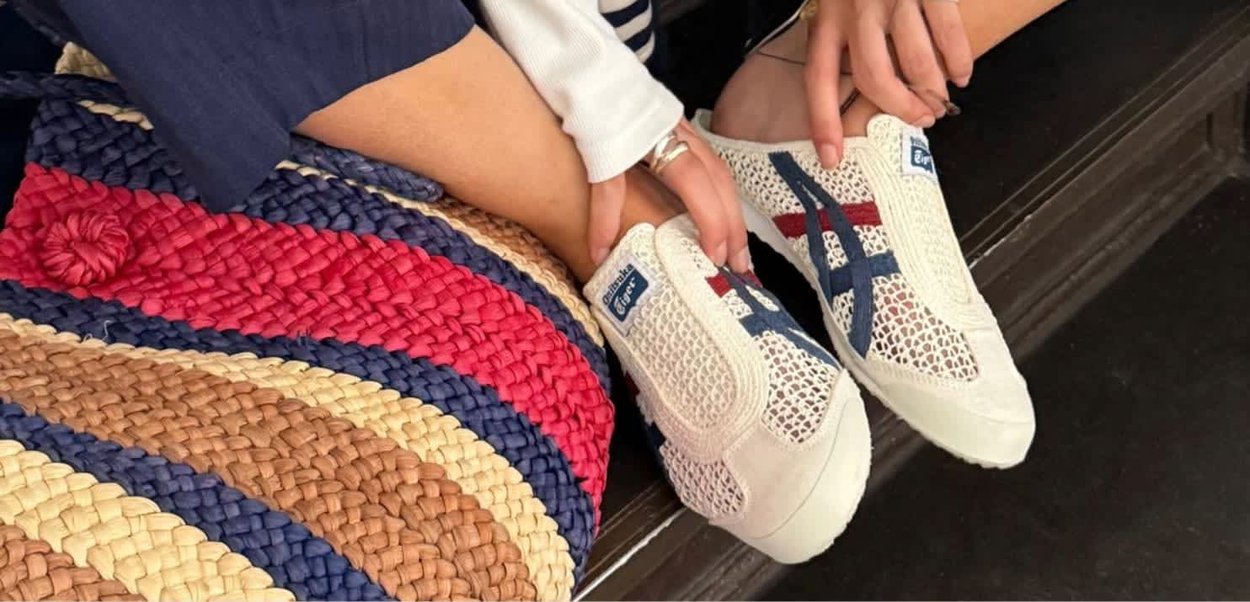 Persona sentada sobre una alfombra tejida multicolor sosteniendo zapatillas blancas tipo slip-on con rayas azules y rojas.