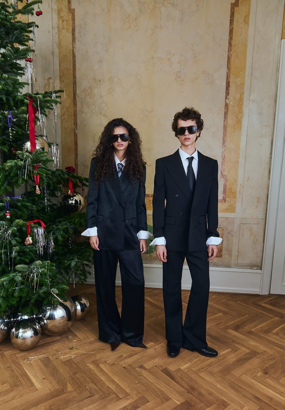 Kaksi henkilöä pukeutuu ylisuuruihin mustiin pukuihin, valkoisiin kauluspaitoihin ja solmittaviin kravattiliin, ja heillä on aurinkolasit päässä. He seisovat koristellun joulukuusen edessä.