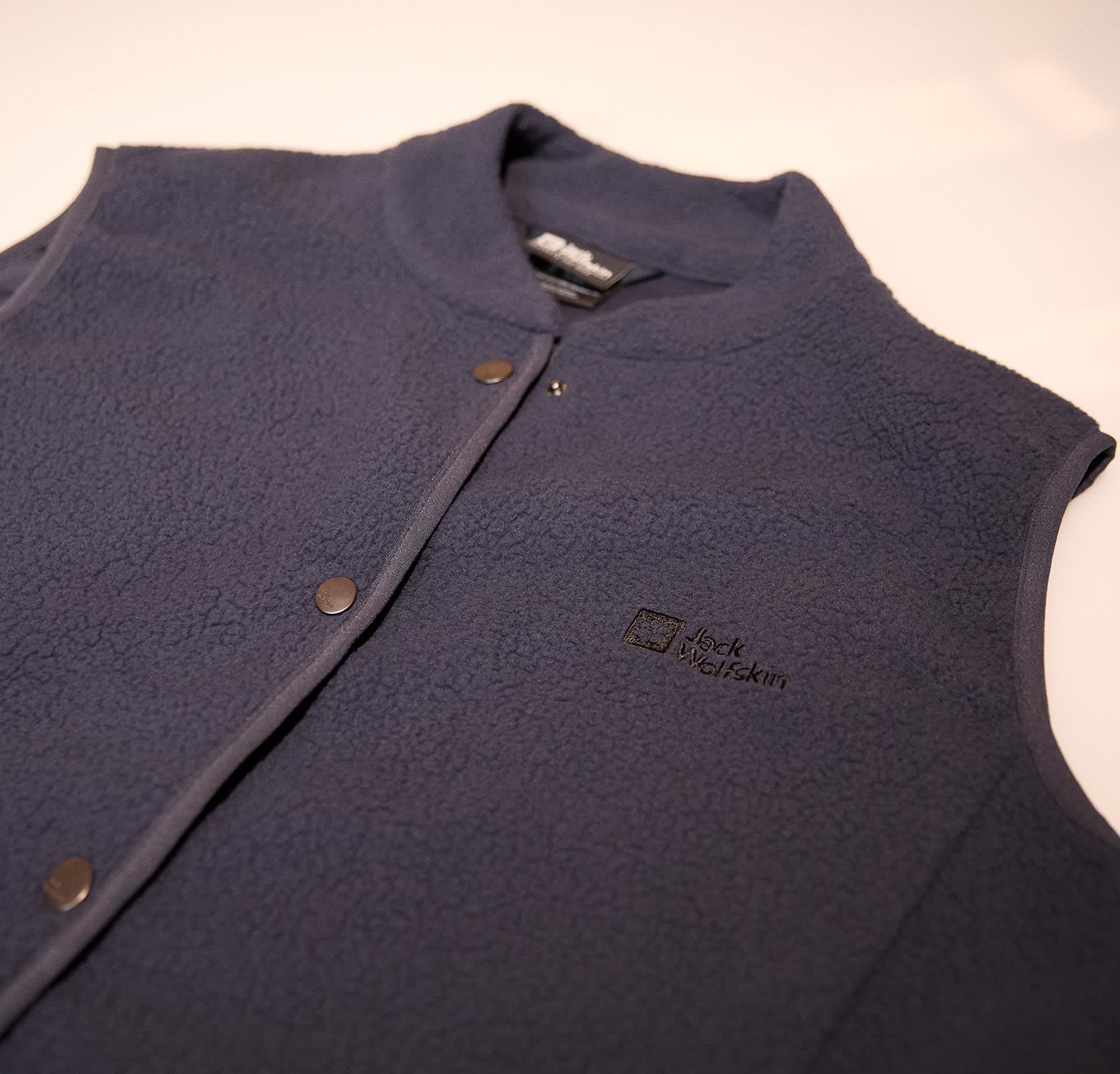 Marinefarbener Fleece-Weste mit gepolstertem Kragen, Druckknöpfen und strukturierter Oberfläche. Das Design beinhaltet ein schwarzes Logo auf der linken Seite.