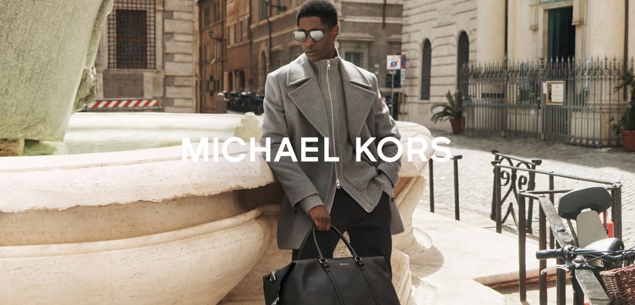 Grå dubbelknäppt rock över en matchande zip-up tröja, kombinerad med en svart läderduffelväska. Släta texturer och moderna designdetaljer.