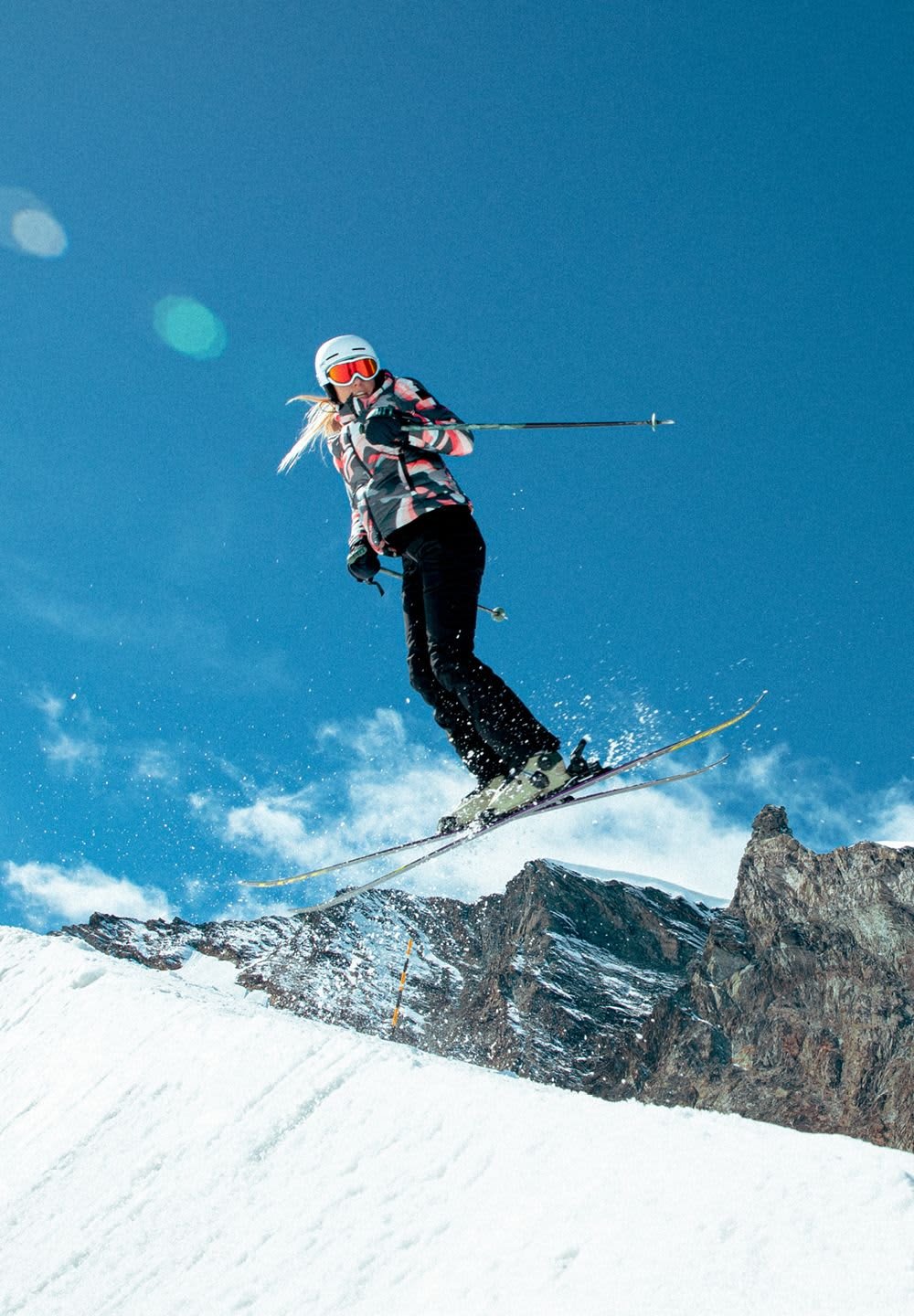 Un schior îmbrăcat într-o jachetă modelată în alb-negru, roz și gri, și pantaloni negri sări de pe o pârtiu acoperită cu zăpadă, cu munți în fundal.