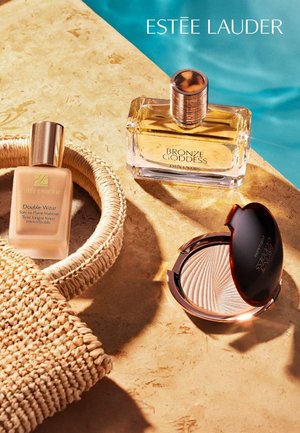 Three Estee Lauder beauty products arranged on a surface: a glass bottle of Bronze Goddess perfume, a foundation in a glass bottle, and a compact highlighter with a shell pattern.