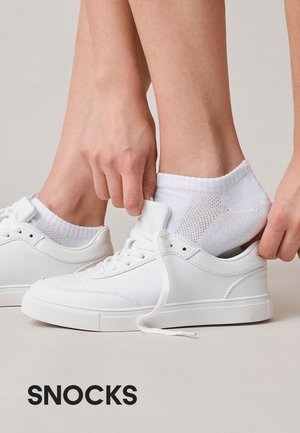 Hænder trækker hvide ankelsokker op over hvide sneakers på bare ben med ordet Snocks nedenfor på en beige baggrund.
