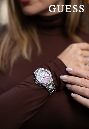 Reloj de acero inoxidable plateado con una esfera de color rosa con patrón y dos subesferas, llevado sobre una camiseta de manga larga marrón. Diseño detallado.