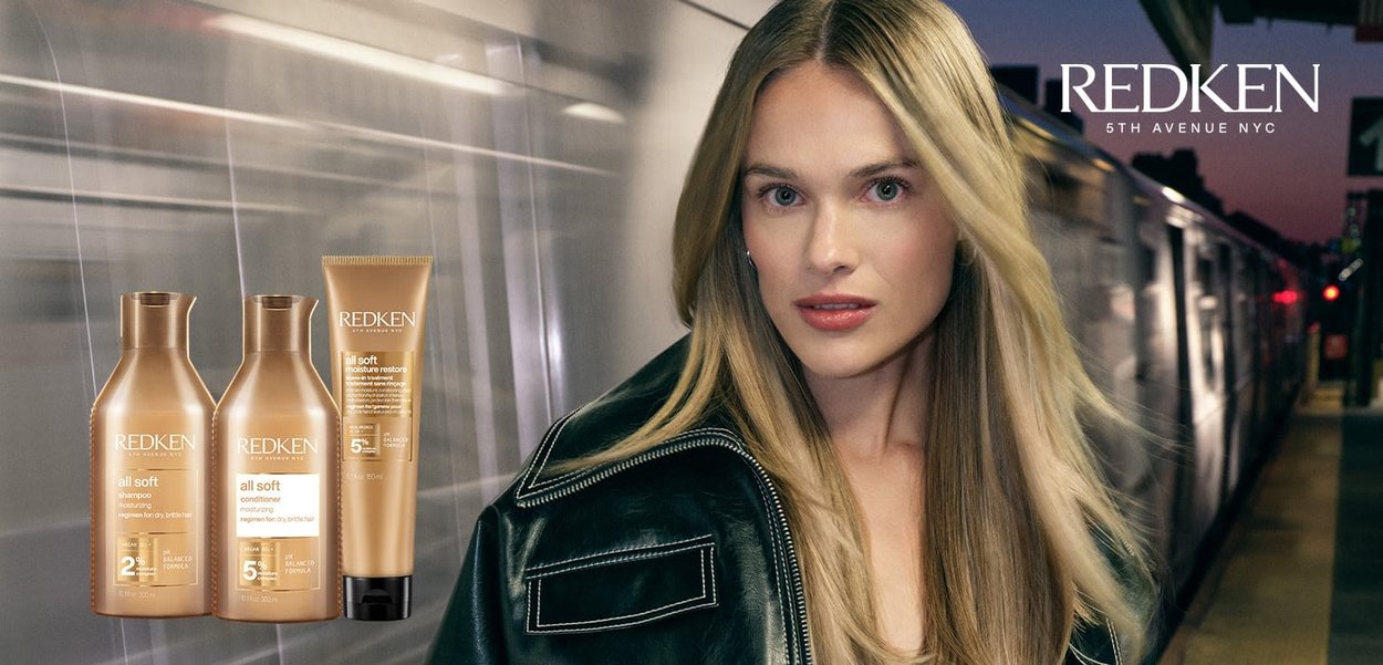 Redken All Soft product line displayed: gold bottles for shampoo and conditioner, and a cream tube. Black leather jacket accent with white stitching.