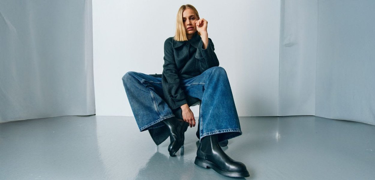 Sorte ankelstøvler med elastiske sidepaneler, båret med en mørk frakke og brede blå denimjeans. Glat tekstur og minimalistisk design, placeret på en reflekterende overflade.