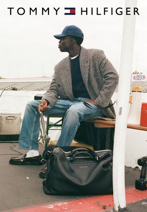 Black leather duffle bag on a concrete surface, accompanied by a man wearing a gray striped coat and blue jeans, seated with tools in hand.