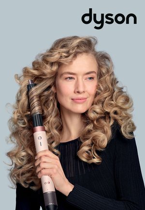Dyson styling tool with a pink grip, black barrel, buttons on the barrel, and a textured dark sweater; model has voluminous curly hair.