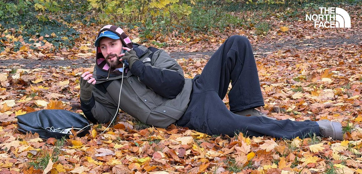 Veste North Face olive avec manches noires, doublure intérieure rose ; pantalon noir ; allongé sur des feuilles orange ; portant des lunettes de soleil ; au téléphone.