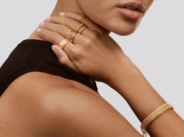 Az arany ékszerek között több gyűrű és egy karkötő található, amelyek textúrázott mintákkal és sima felületekkel rendelkeznek, egy csupasz karon és kézen viselve.