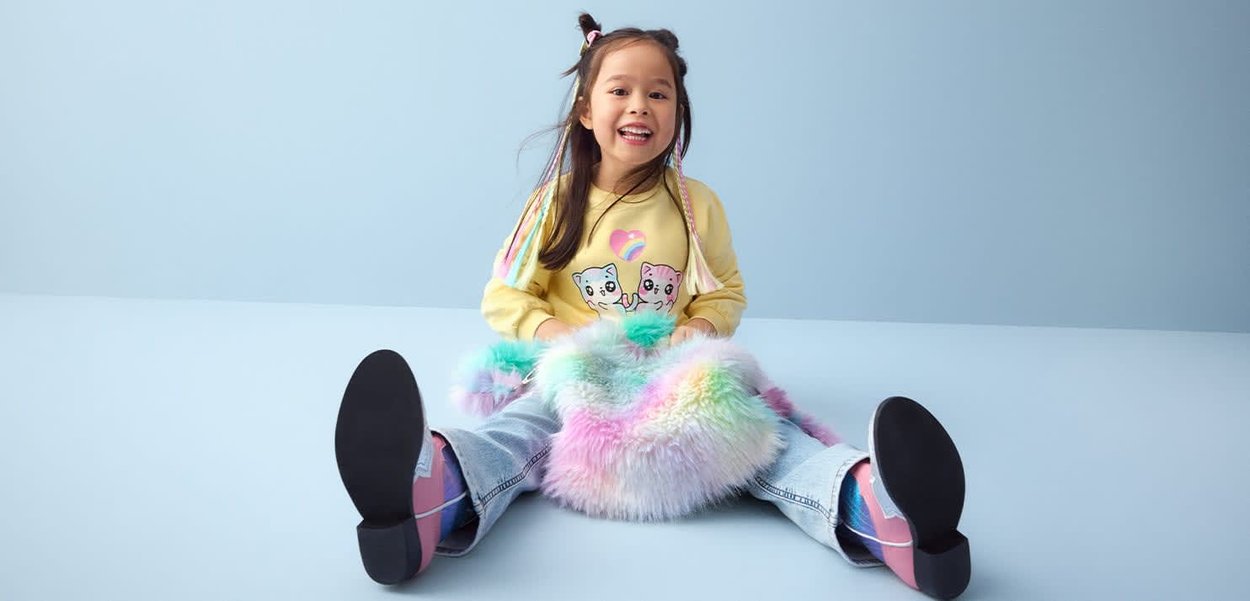 Jong meisje met kleurrijke haren zit op de vloer, lacht en houdt een pluizige pastel regenboogtas vast, draagt een gele trui met hondenprints.