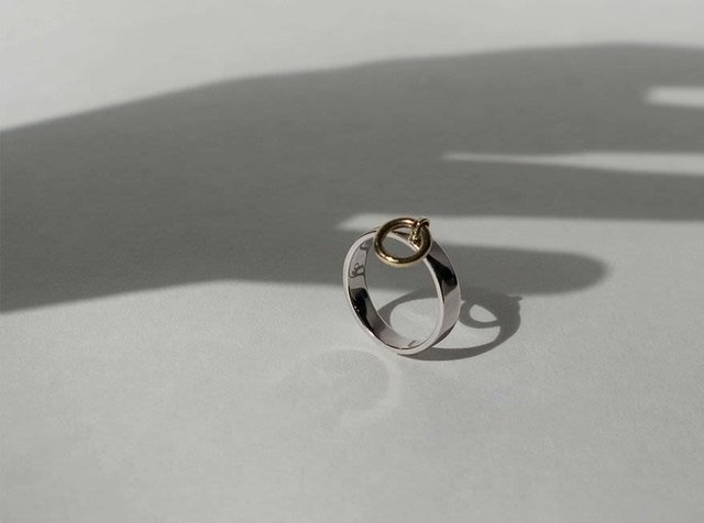 Ez egy ezüst gyűrű, sima felülettel, amelyen egy kis arany karika függ, lágy árnyékot vetve egy világos felületre.