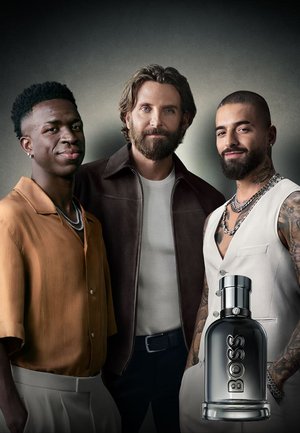 Trois hommes se tiennent ensemble, portant une veste marron, une chemise beige et un pantalon, et un gilet blanc. Une bouteille de parfum argentée avec "BOSS" est devant eux.