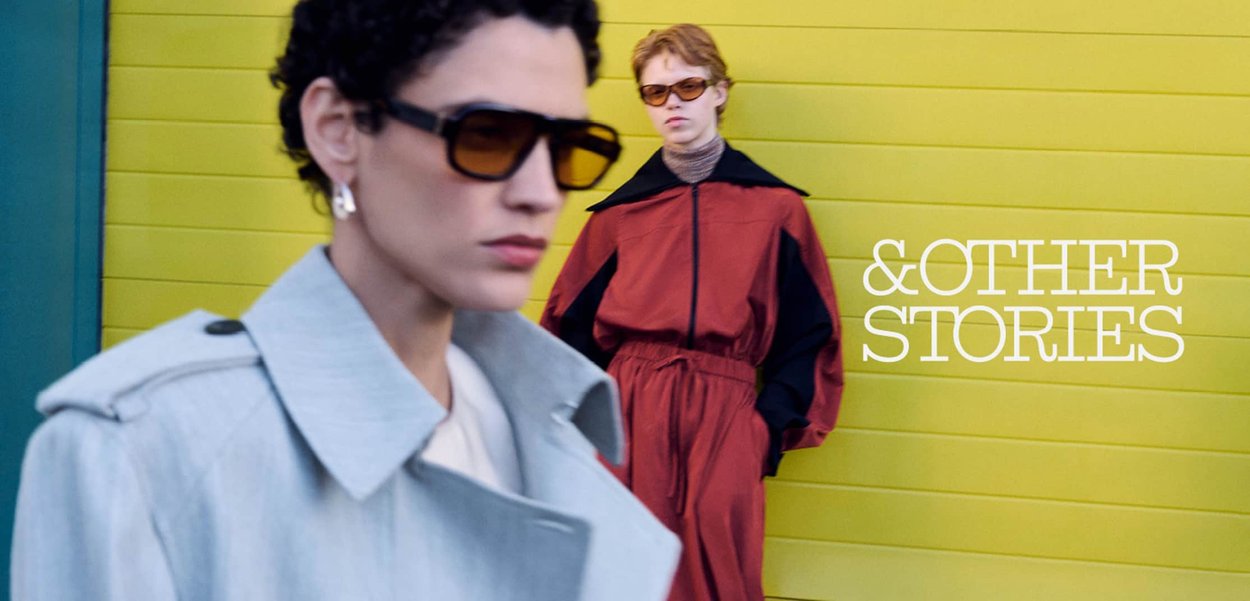 Twee modieuze modellen dragen een zonnebril en stijlvolle jassen en staan voor een gele muur met de tekst "&OTHER STORIES".