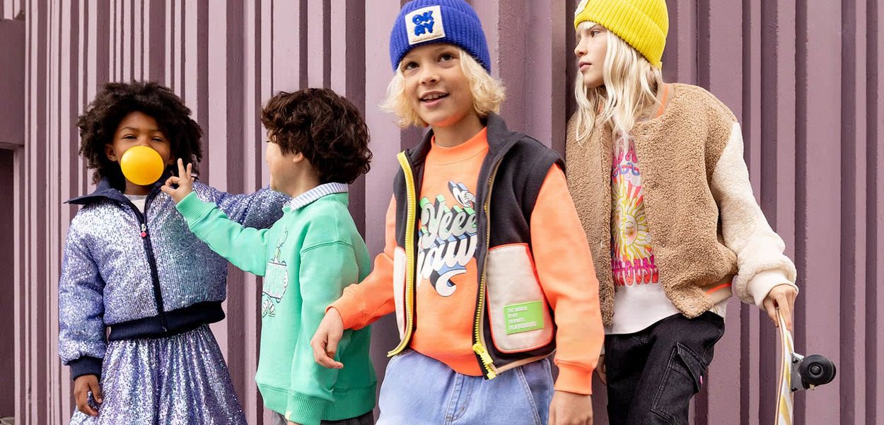 Vier Kinder tragen farbenfrohe, strukturierte Outfits: eine mit Pailletten besetzte Jacke, einen Fleece-Bomber, ein grafisches Sweatshirt und lebhafte Mützen, ergänzt durch verspielte Accessoires.