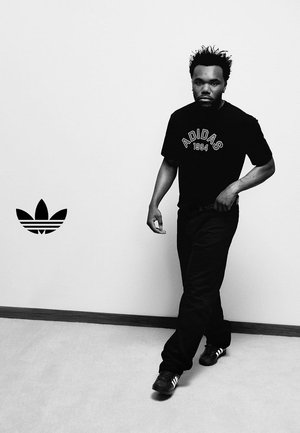 Mężczyzna idący wewnątrz budynku, ubrany w czarną koszulkę Adidas z napisem "1994", czarne spodnie i czarne buty sportowe Adidas, z logo Adidas na pobliskiej białej ścianie.