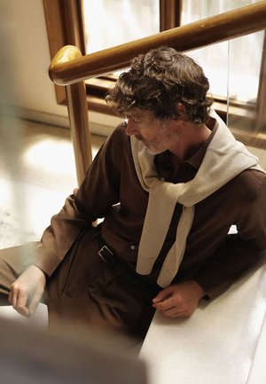 Una camicia marrone a bottoni indossata sotto un maglione beige chiaro appoggiato attorno al collo, in contrasto con pantaloni marroni scuri, seduta su marmo.