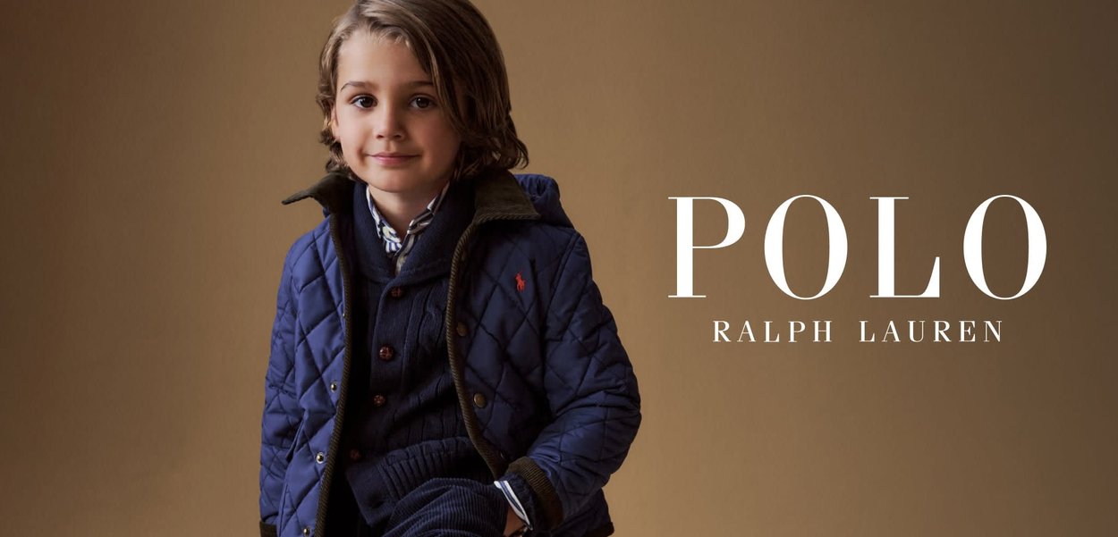 Niño pequeño sentado en un taburete de madera, vestido con chaqueta acolchada azul marino, suéter y pantalones de pana, contra un fondo beige.