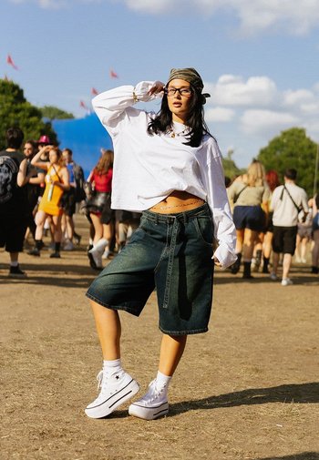 Denim shorts, loose fit, dark wash, paired with a white cropped long-sleeve shirt and white platform sneakers. Background with festival crowds.