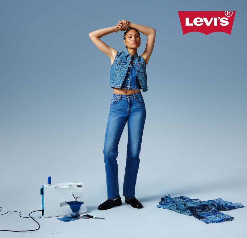 Persoon in spijkerjasje en jeans staat tussen een naaimachine met stof en een stapel spijkerpatches, met het Levi's-logo erboven.