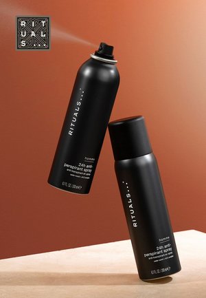 Des sprays anti-transpirants aérosols noirs et sticks avec des accents argentés, étiquetés "RITUALS", sur un fond orange sur une surface beige.