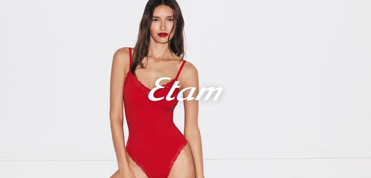 Femme aux longs cheveux bruns portant un body rouge orné de dentelle, debout devant un fond blanc uni.