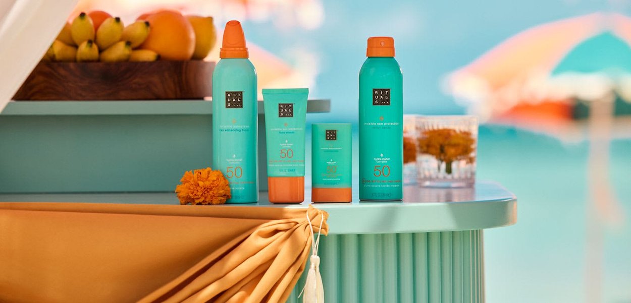 Cuatro botellas turquesas de protector solar con tapones naranjas etiquetados con SPF 50 sobre una mesa azul junto a una piscina con un cuenco de frutas al fondo.