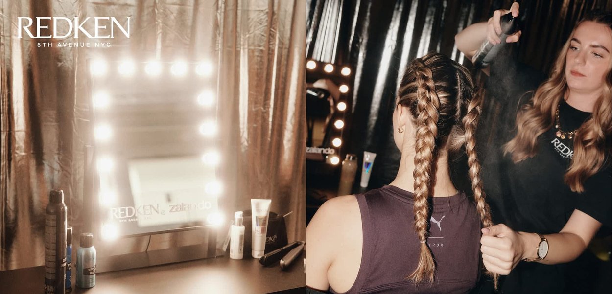 Schminkspiegel mit hellen Glühbirnen neben Haarpflegeprodukten. Haare in zwei Zöpfen gestylt; Stylistin sprüht Haarspray auf. Der Hintergrund zeigt glänzende schwarze Vorhänge.