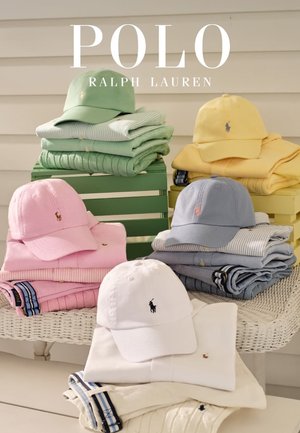 Conjuntos apilados de ropa Polo Ralph Lauren en colores pastel con gorras a juego sobre una mesa de mimbre blanca y cajas de madera.