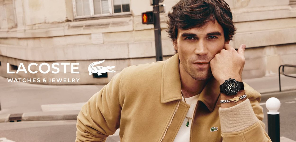 Mann in beiger Jacke zeigt schwarze Uhr und silbernes Armband, steht auf einer Stadtstraße mit Fußgängerampel im Hintergrund.