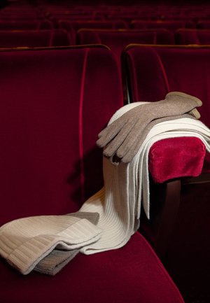 Braune Handschuhe auf einem hellgrauen, gerippten Schal, der auf einem burgunderroten Theatersitz liegt. Der Schal hat gefaltete Enden mit kontrastierenden Farben.