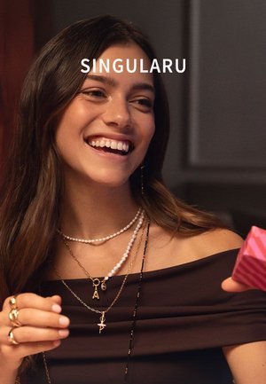 Top marrone con spalle scoperte e collane dorate e di perle a strati. Gli accessori includono un ciondolo a forma di lettera e un oggetto rosa dettagliato in mano.