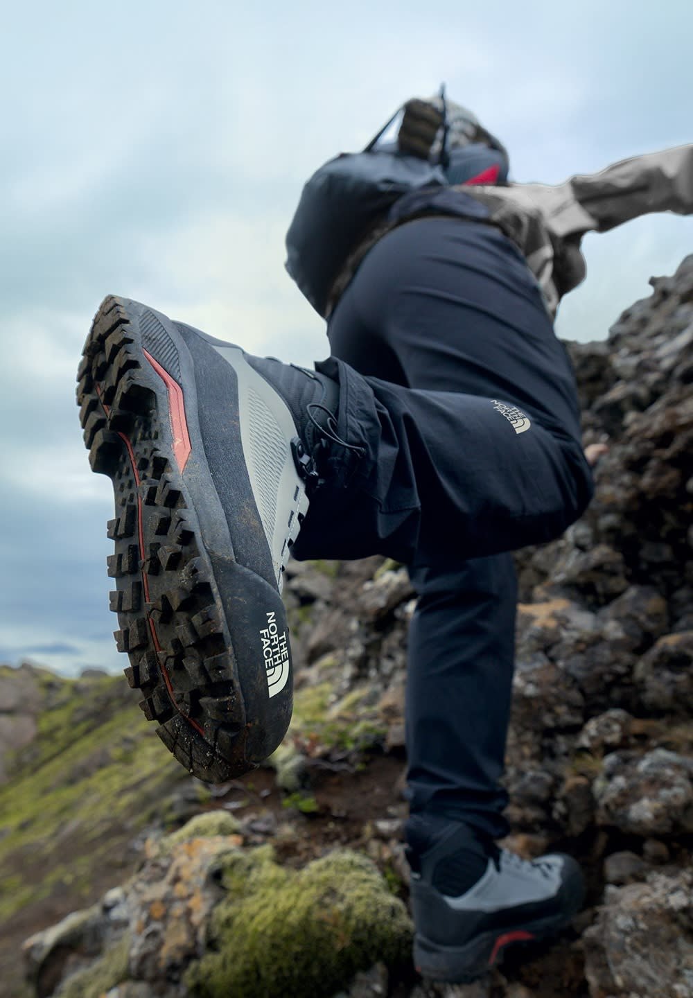 Scarponi da trekking grigi e neri con suola robusta, caratterizzati da tasselli profondi e dettagli rossi, posati su un terreno roccioso ricoperto di muschio.