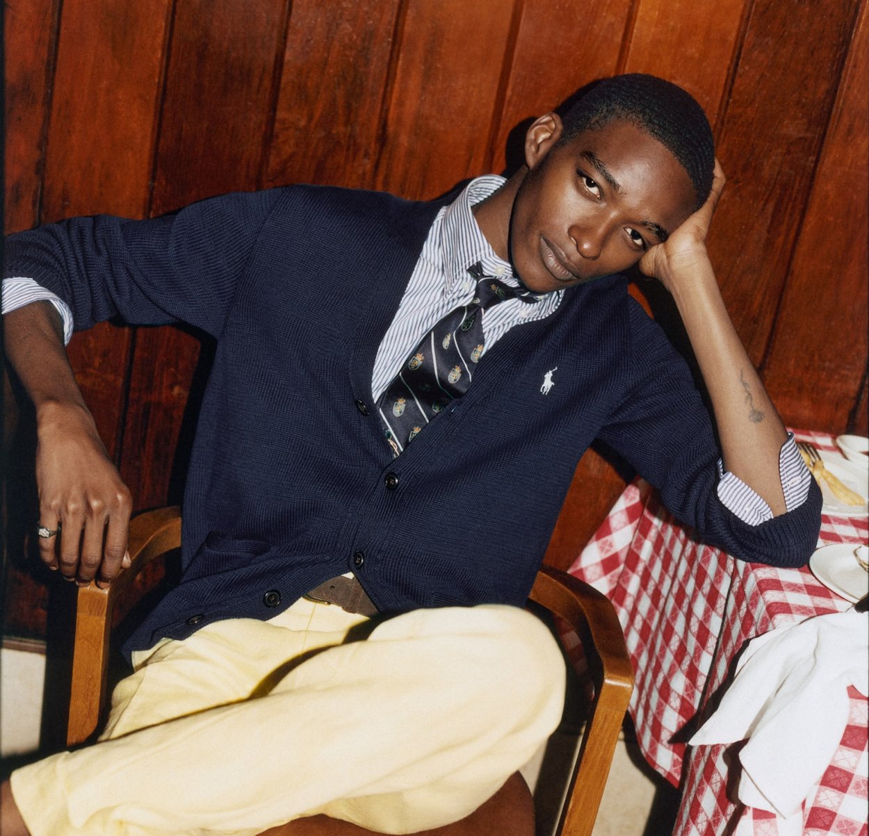 Jonge man gekleed in een marineblauwe cardigan, gestreept shirt en een stropdas met patroon, ontspannen zittend met één arm op een houten stoel en de andere ter ondersteuning van zijn hoofd.