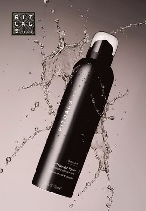 Bouteille cylindrique noire avec une forme courbée, capuchon transparent. Le texte indique "RITUALS," "mousse de douche," et ingrédients. Des éclaboussures d'eau entourent la bouteille.